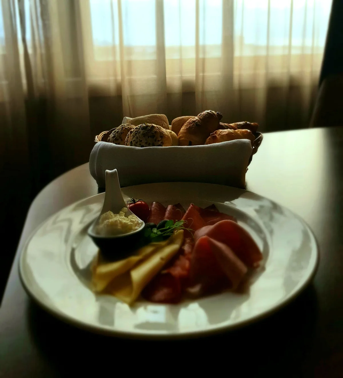 Bread and a plate of avocado, tomato and cheese beside a softly lit window